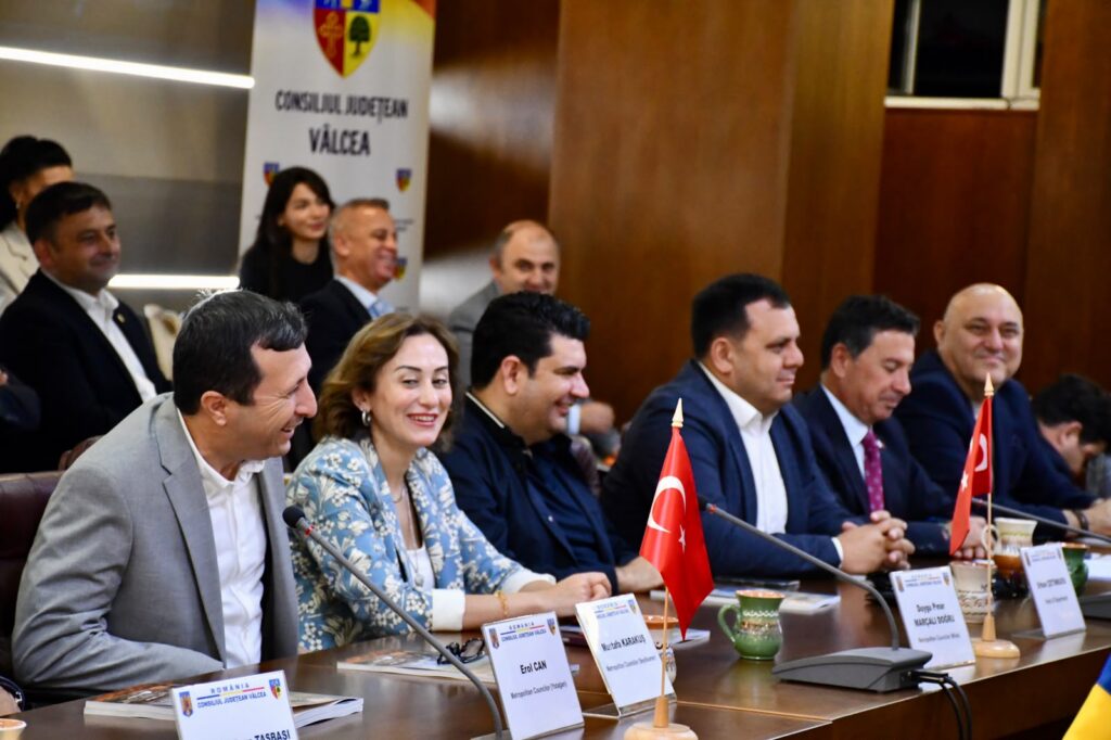 Group of officials seated at a long conference table, with small Turkish flags, nameplates, and mugs in front of them during a formal meeting.