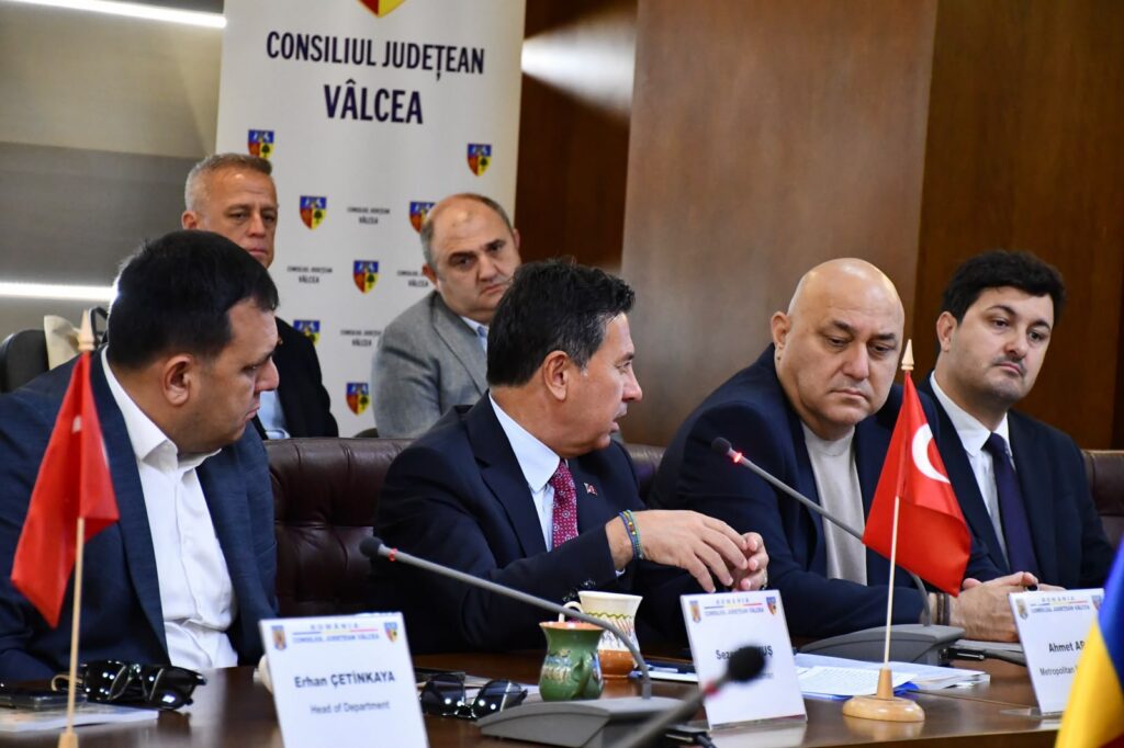 Group of men in suits at a council meeting; Turkish flag and name placards on the table, banner behind reads Consiliul Judetean Valcea