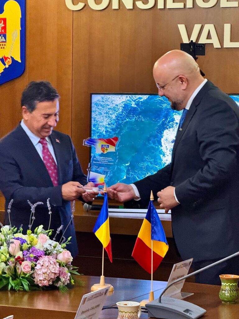 Two men in suits exchange a small ceremonial item during a formal meeting, with Romanian and Moldovan flags on the table and flowers nearby.