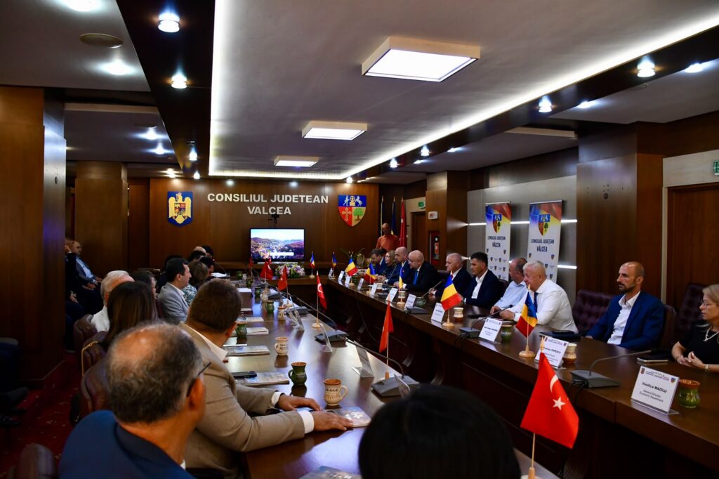 Officials seated around a long conference table in a government meeting room, with Romanian flags and the Valcea County Council crest on the wall.