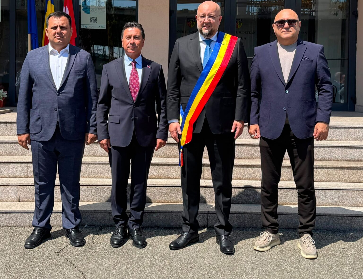 Four men in suits pose on stone steps outside a building; the third man wears a blue-yellow-red ceremonial sash.