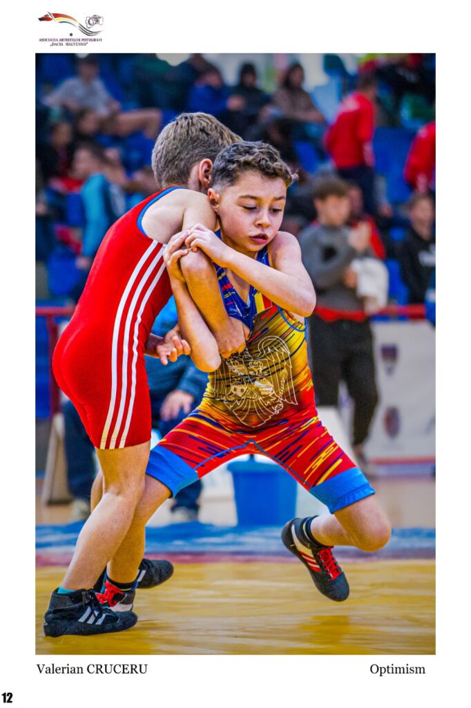 Two young wrestlers grapple in a gym during a match, one in red, the other in a colorful singlet, focused and determined in front of spectators.