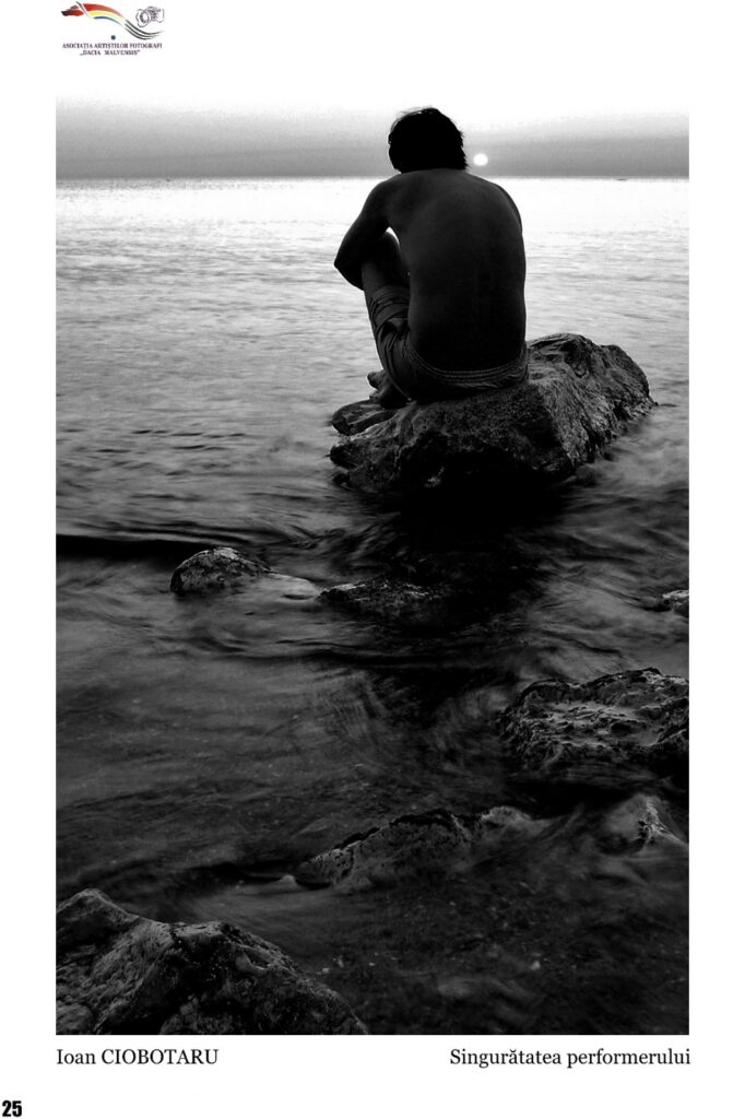 Man sits on a rock at the water's edge, back to the camera, watching a sunset over the sea (black and white).