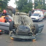Emergency responders attend to a overturned car on a residential street; ambulance and fire truck parked nearby.