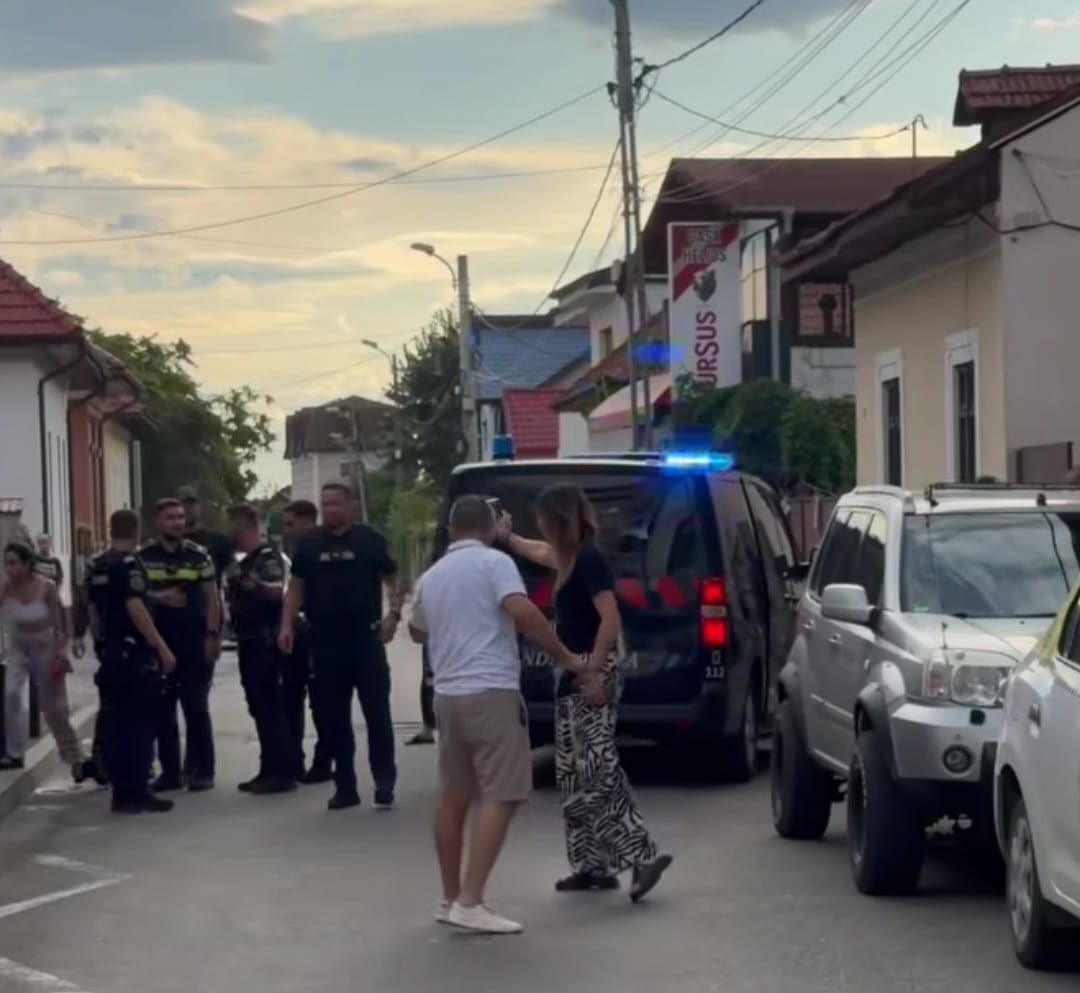 Group of police officers and civilians stand on a residential street beside a police SUV with blue lights on.