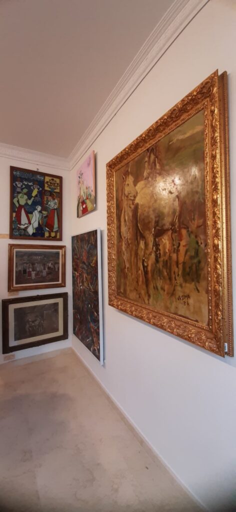 WhatsApp Image 2026-07-11 at 224051 - Jurnalul Olteniei – Știri din Craiova și Oltenia Corner view of an art gallery wall with several framed paintings; the largest on the right is a gold ornate frame with a desert scene featuring camels.
