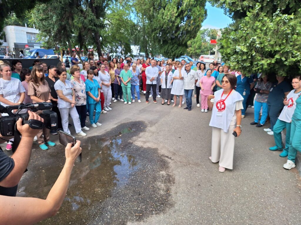 WhatsApp Image 2026-07-20 at 110652 - Jurnalul Olteniei – Știri din Craiova și Oltenia Large group of medical professionals in scrubs and white coats gathered on a street outdoors, with cameras recording nearby actors in the foreground.