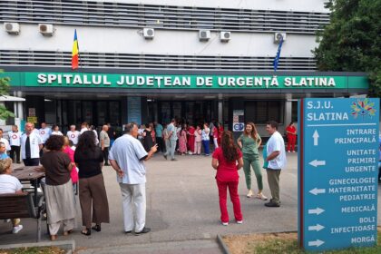 Hospital entrance with a crowd of people outside, Romanian and EU flags above the green sign reading 'Spitalul Judetean de Urgență Slatina' (Slatina Emergency County Hospital). In the foreground, medical staff in white coats mingle near a blue sign with arrows.