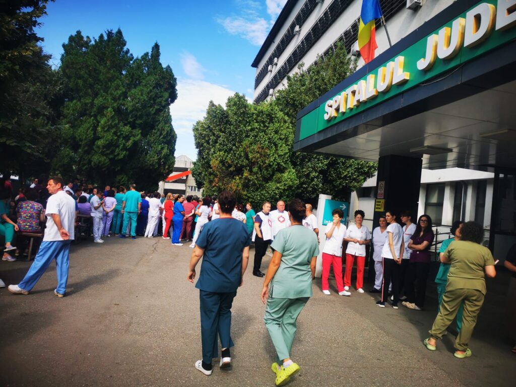 WhatsApp Image 2026-07-20 at 110755 - Jurnalul Olteniei – Știri din Craiova și Oltenia Crowd of hospital staff in colorful scrubs and uniforms gathered outside a hospital entrance with a Romanian flag overhead.