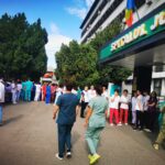 Crowd of hospital staff in colorful scrubs and uniforms gathered outside a hospital entrance with a Romanian flag overhead.