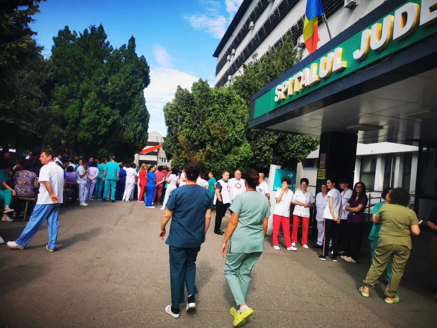 WhatsApp Image 2026-07-20 at 110755 - Jurnalul Olteniei – Știri din Craiova și Oltenia Crowd of hospital staff in colorful scrubs and uniforms gathered outside a hospital entrance with a Romanian flag overhead.