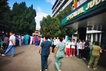 Crowd of hospital staff in colorful scrubs and uniforms gathered outside a hospital entrance with a Romanian flag overhead.
