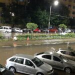 Nighttime street scene with a flooded road and parked cars along the curb; water covers the roadway in a city