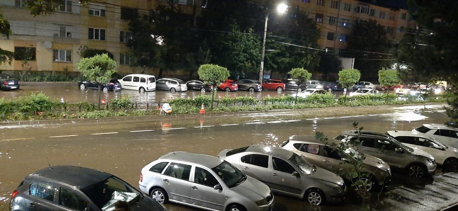 WhatsApp Image 2026-07-21 at 221045 - Jurnalul Olteniei – Știri din Craiova și Oltenia Nighttime street scene with a flooded road and parked cars along the curb; water covers the roadway in a city