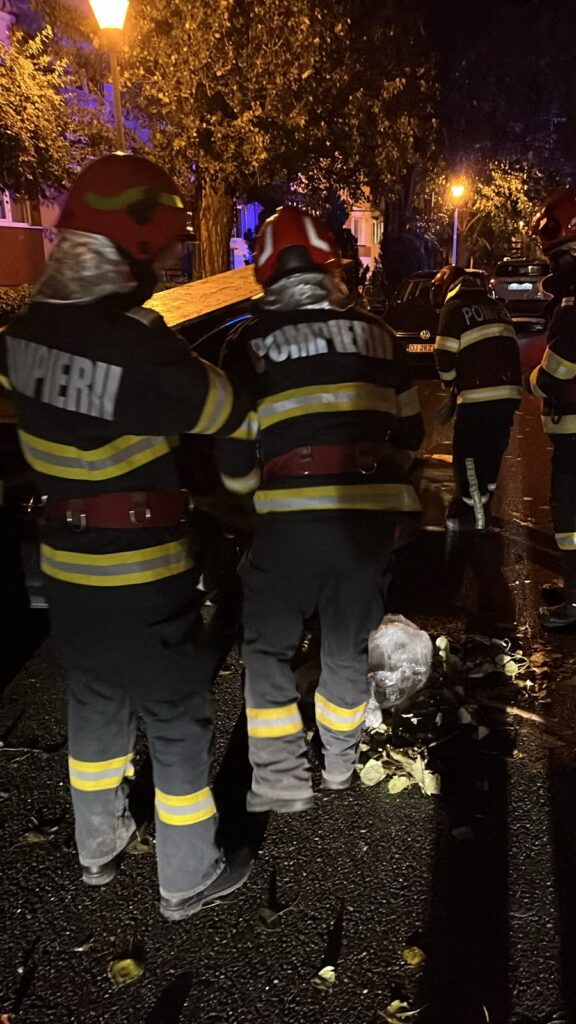 WhatsApp Image 2026-07-21 at 230919 1 - Jurnalul Olteniei – Știri din Craiova și Oltenia Night-time scene of firefighters in reflective gear and red helmets tending to debris on a wet street with leaves and a parked car nearby, illuminated by street lamps.
