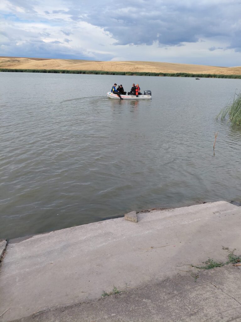 WhatsApp Image 2026-07-25 at 102514 1 - Jurnalul Olteniei – Știri din Craiova și Oltenia Group of people in a small inflatable boat wearing life jackets on a calm lake, with a grassy shoreline and distant sandy hills under a cloudy sky.