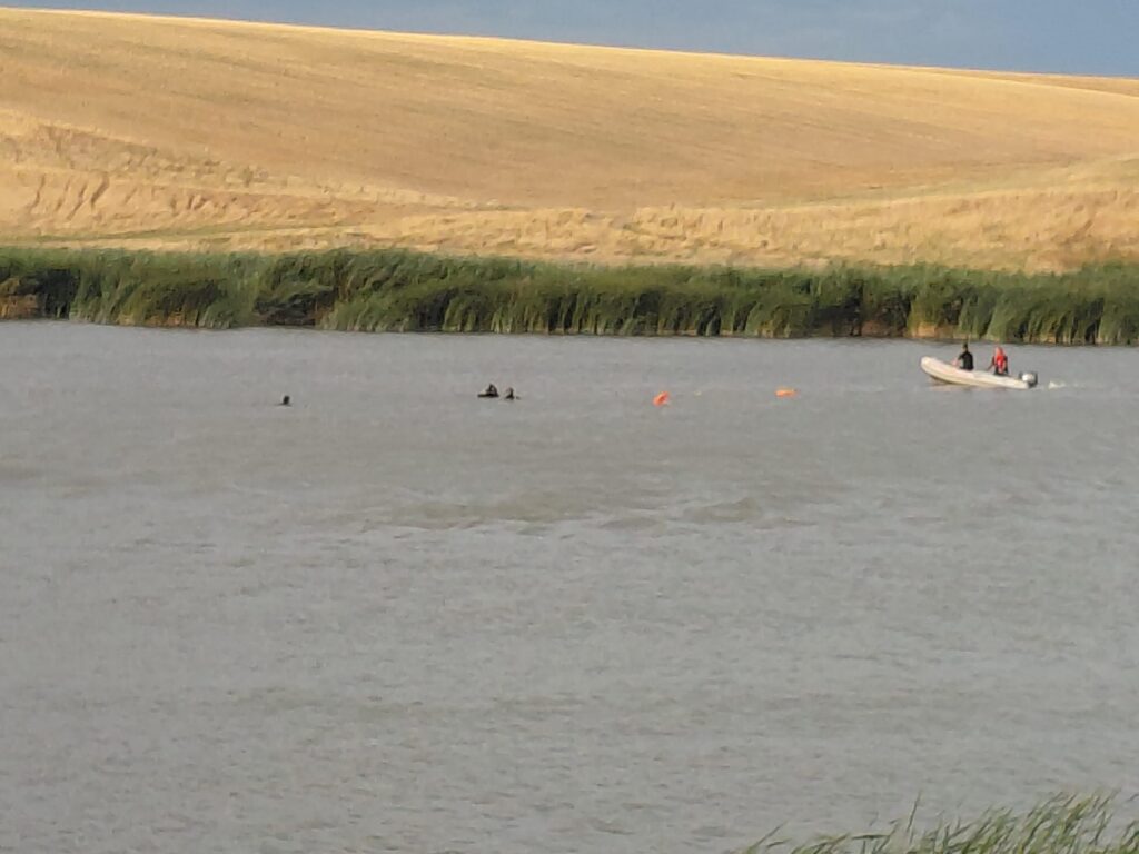 WhatsApp Image 2026-07-25 at 102514 - Jurnalul Olteniei – Știri din Craiova și Oltenia A calm river with a small white boat carrying two people near the grassy bank, a sandy hill in the background and orange buoys in the water.
