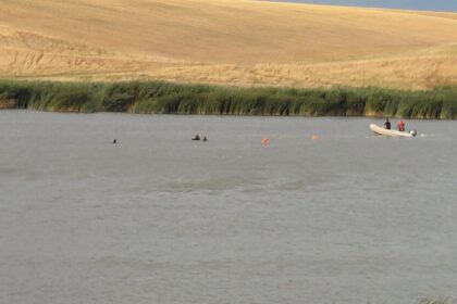 A calm river with a small white boat carrying two people near the grassy bank, a sandy hill in the background and orange buoys in the water.