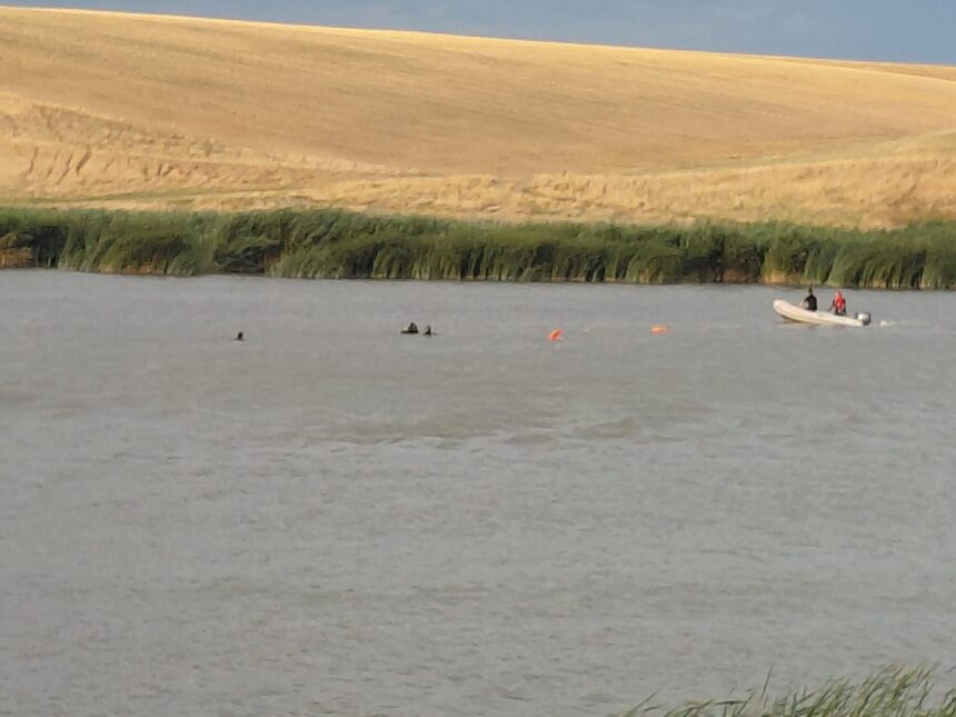 A calm river with a small white boat carrying two people near the grassy bank, a sandy hill in the background and orange buoys in the water.