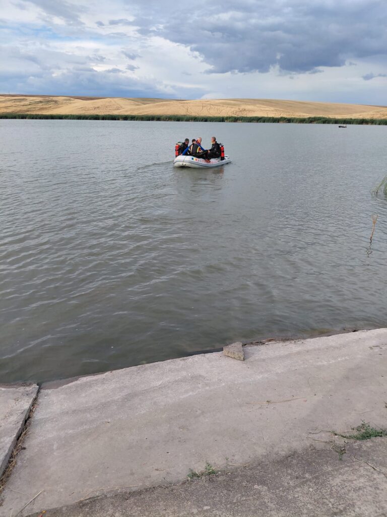 WhatsApp Image 2026-07-25 at 102602 1 - Jurnalul Olteniei – Știri din Craiova și Oltenia A small inflatable boat carrying four people wearing life jackets on a calm lake, with sandy hills and cloudy sky in the background.