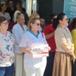 A diverse crowd of healthcare workers and civilians standing outdoors near a hospital entrance, some wearing scrubs and badges.