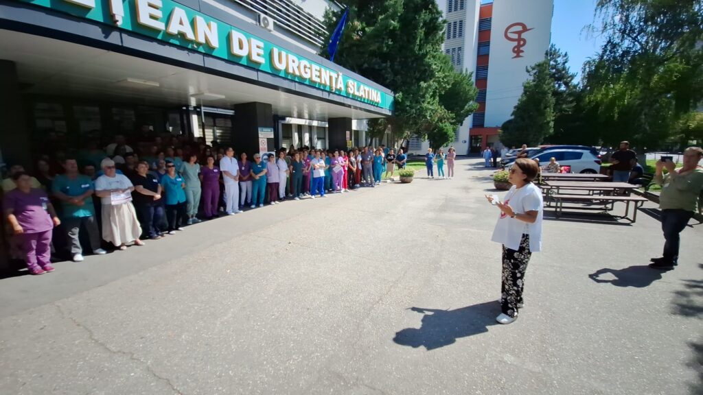 A line of healthcare workers and staff stand outside a hospital entrance while a woman speaks to them from the foreground.