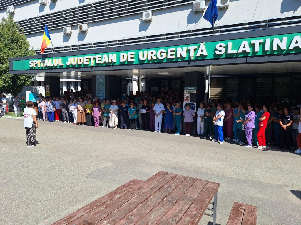 Long line of healthcare workers in scrubs and casual clothes outside a hospital entrance, Romanian flag overhead on a sunny day.
