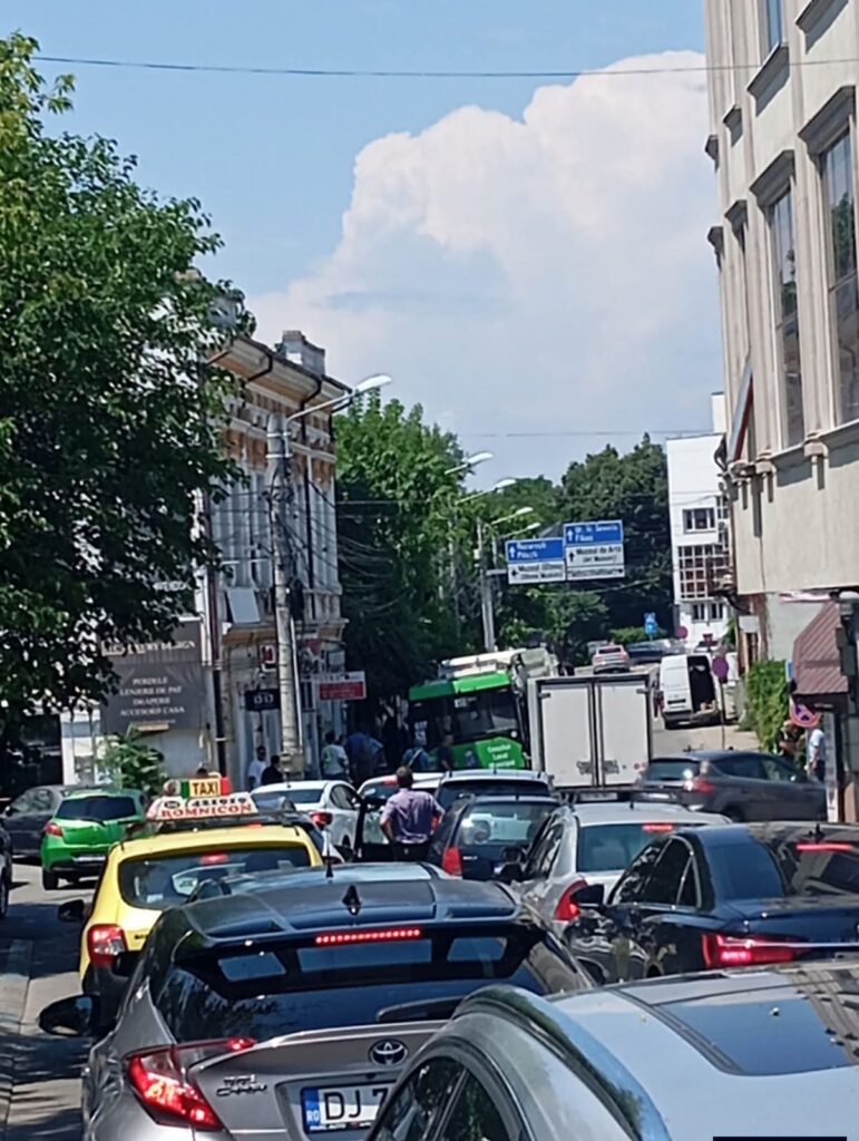 asfalt autobuz WhatsApp Image 2026-07-03 at 142740 - Jurnalul Olteniei – Știri din Craiova și Oltenia Busy city street with cars and a green bus, tall buildings on both sides, trees, and a blue sky with clouds.