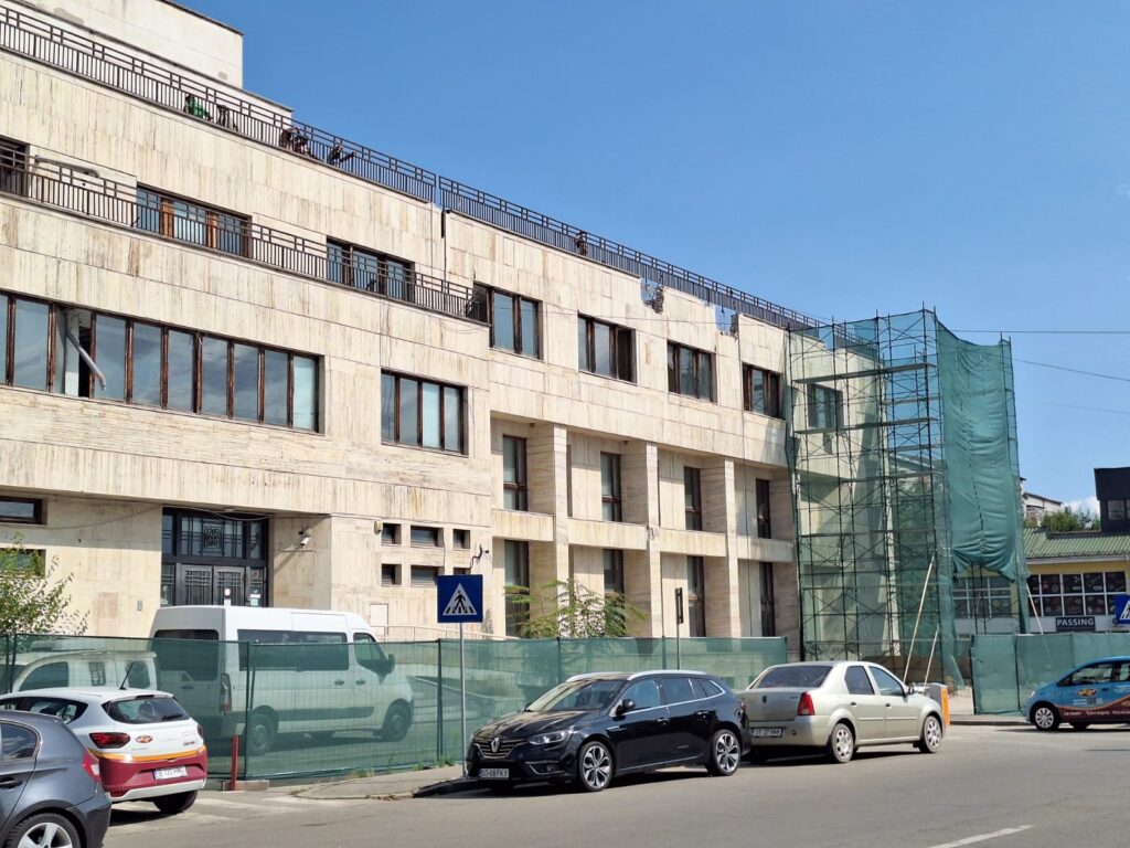 bcr slatina victoria intermed 1 - Jurnalul Olteniei – Știri din Craiova și Oltenia Construction site next to a beige stone building; scaffolding with green netting on the right, blue sky above, street with parked cars and a pedestrian crossing sign