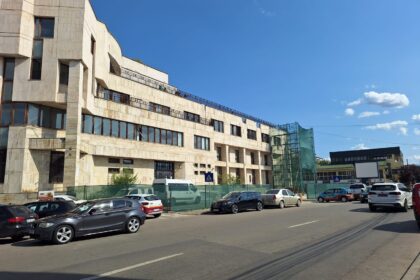 Beige multi-story building under construction with scaffolding and green netting, parked cars along the street, under a clear blue sky.