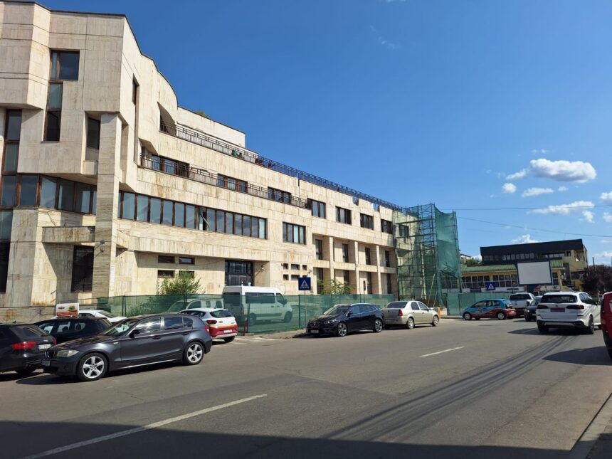Beige multi-story building under construction with scaffolding and green netting, parked cars along the street, under a clear blue sky.
