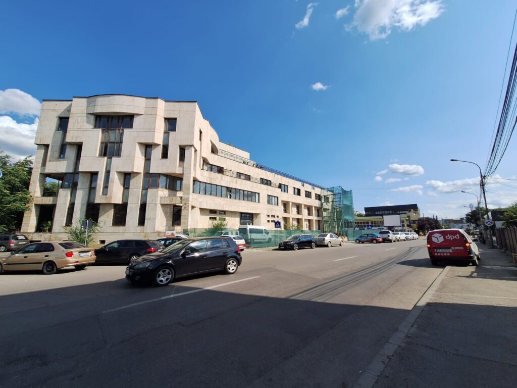 bcr slatina victoria intermed 3 - Jurnalul Olteniei – Știri din Craiova și Oltenia Modern beige stone building with staggered windows under construction along a busy street, cars parked in front, blue sky above, daytime.