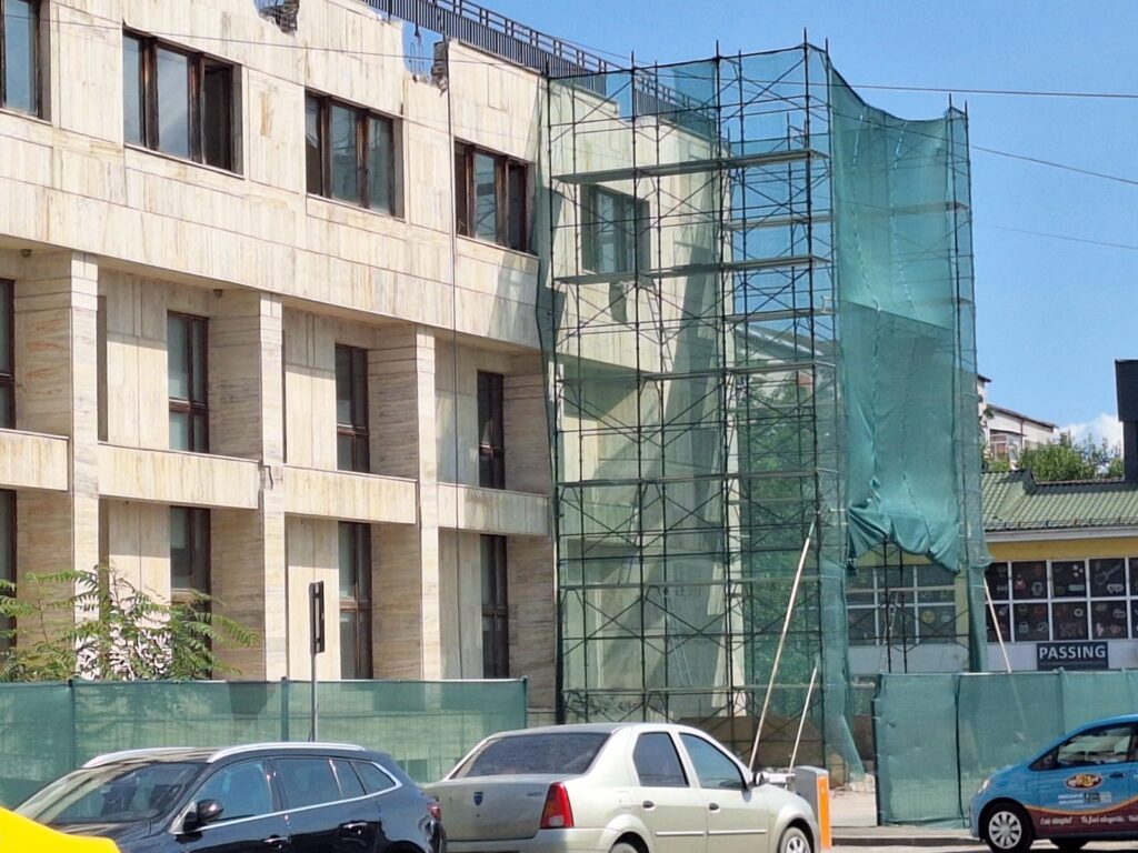 bcr slatina victoria intermed 4 - Jurnalul Olteniei – Știri din Craiova și Oltenia Construction scaffolding wrapped in green mesh beside a beige stone building under renovation. Cars parked in front.