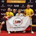 Four men pose on a competition stage with medals; two wear yellow shirts, one in white, holding a banner that says '77 Laboratory MMA - BJJ'.