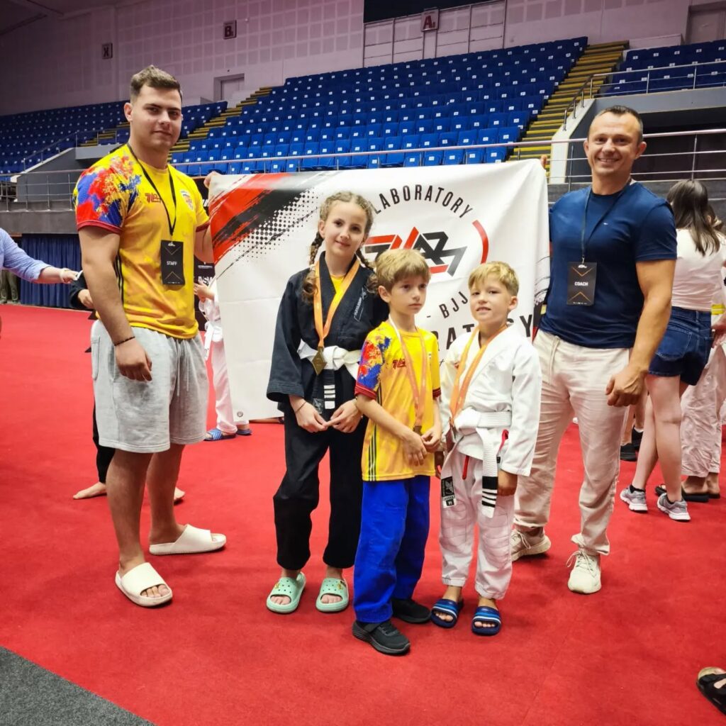 Group of two kids in martial arts uniforms with medals posing with two coaches on a red competition mat.