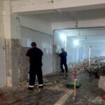 Room under renovation with tiled walls, exposed plaster, and rubble on the floor; two workers stand overseeing the demolition.