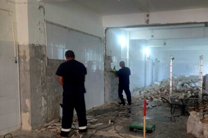 Room under renovation with tiled walls, exposed plaster, and rubble on the floor; two workers stand overseeing the demolition.