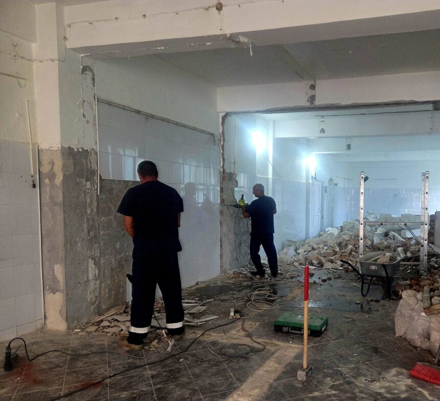 Room under renovation with tiled walls, exposed plaster, and rubble on the floor; two workers stand overseeing the demolition.