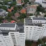 Drone view of a dense urban area with a tall white apartment building in the foreground and a mix of houses, trees, and streets beyond.