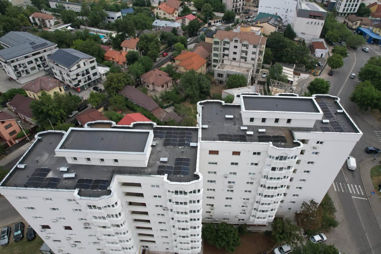 Drone view of a dense urban area with a tall white apartment building in the foreground and a mix of houses, trees, and streets beyond.