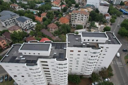 Drone view of a dense urban area with a tall white apartment building in the foreground and a mix of houses, trees, and streets beyond.