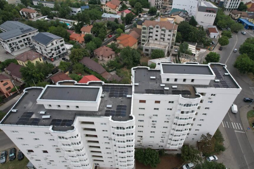 Drone view of a dense urban area with a tall white apartment building in the foreground and a mix of houses, trees, and streets beyond.