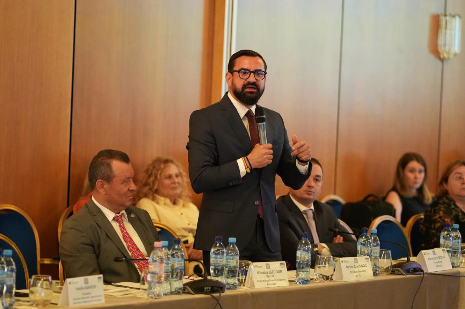 Man in a dark suit stands and speaks into a handheld microphone at a panel discussion, with seated colleagues nearby.