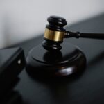 Judge's gavel with a gold band rests on a black desk, ready to strike a sound block in a courtroom setting.