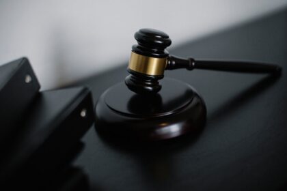 Judge's gavel with a gold band rests on a black desk, ready to strike a sound block in a courtroom setting.