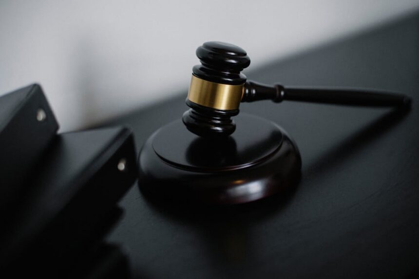 Judge's gavel with a gold band rests on a black desk, ready to strike a sound block in a courtroom setting.