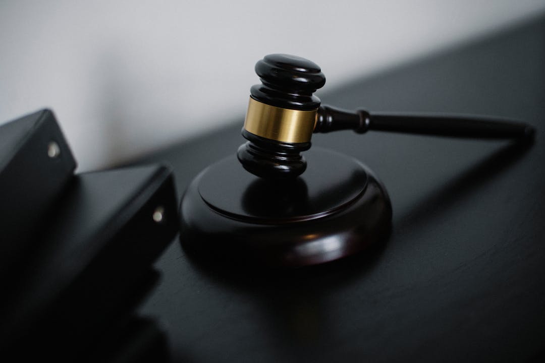 Judge's gavel with a gold band rests on a black desk, ready to strike a sound block in a courtroom setting.