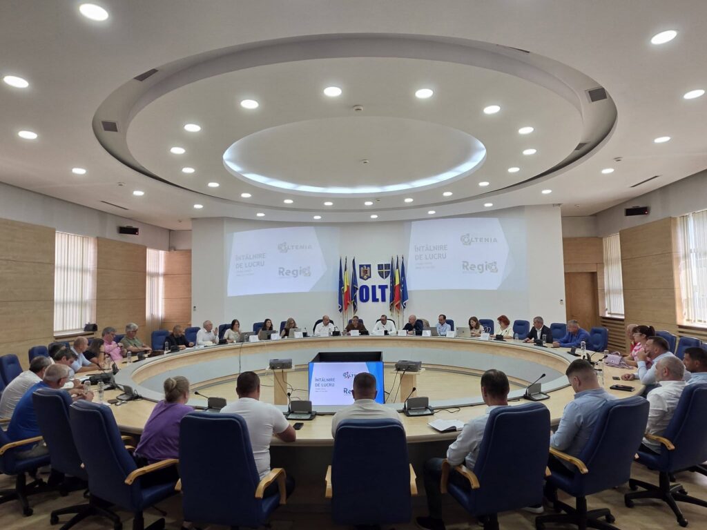 Delegates seated around a large oval conference table in a formal meeting room with flags and projection screens in the background.