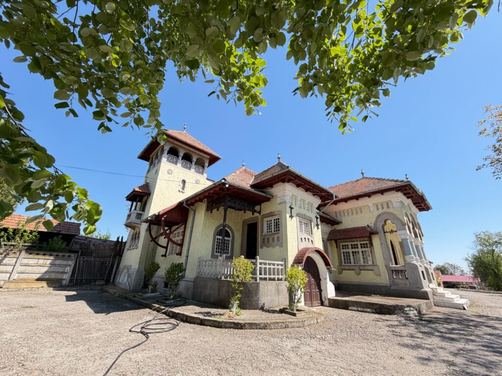 conacul romano plesoiu 3 - Jurnalul Olteniei – Știri din Craiova și Oltenia Villa with ornate cream facade and red-tiled roof seen through leafy branches on a sunny day