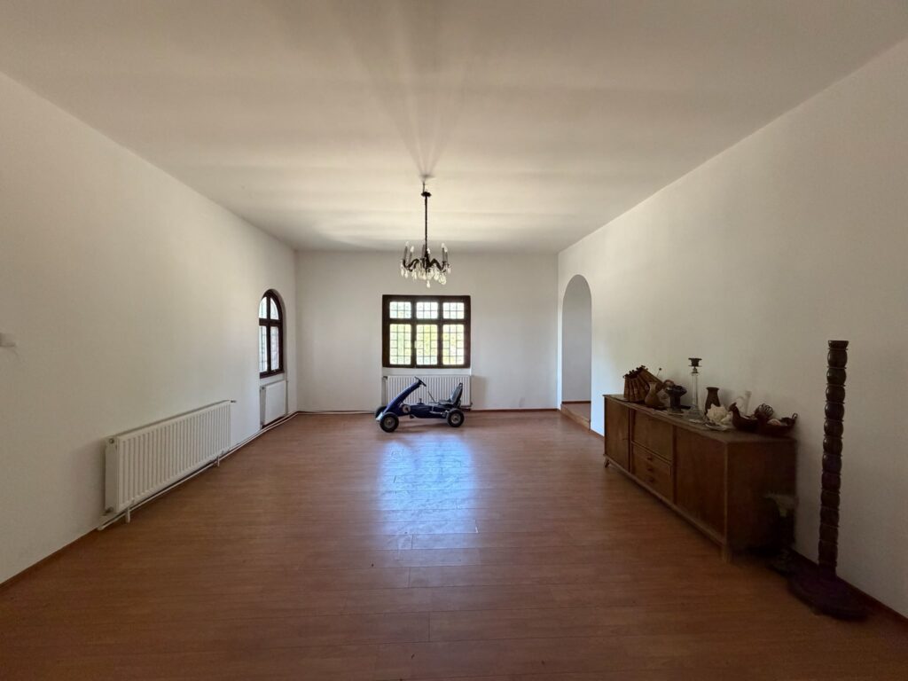 conacul romano plesoiu 6 - Jurnalul Olteniei – Știri din Craiova și Oltenia Bright, sparsely furnished living room with a hanging chandelier, wooden floor, and a blue ride-on toy car centered.